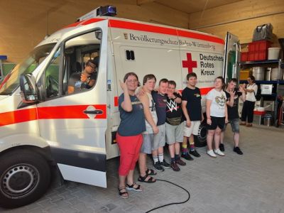 Die Teilnehmenden des Erste-Hilfe-Kurses stehen nebeneinander aufgereiht neben einem Rettungswagen, welcher in einer Halle steht..